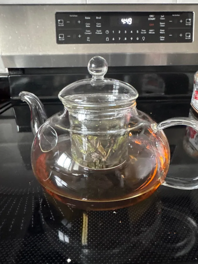 The glass teapot full of amber tea with leaves in the insert, standing on a cooker top. Photographed by a***r, a verified buyer