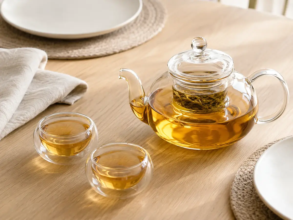 A glass teapot of amber tea on a light oak table laid for two, with two small clear glass cups poured beside it